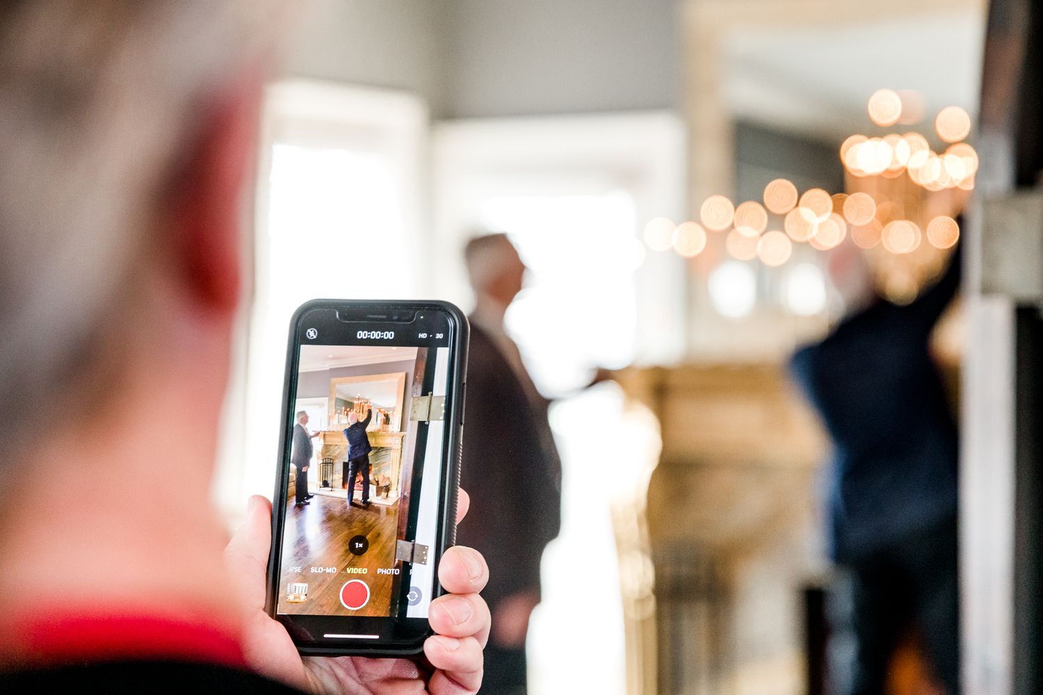 Person taking photo on smartphone of wedding ceremony with bokeh lights in background by Stephanie Coulter Photography
