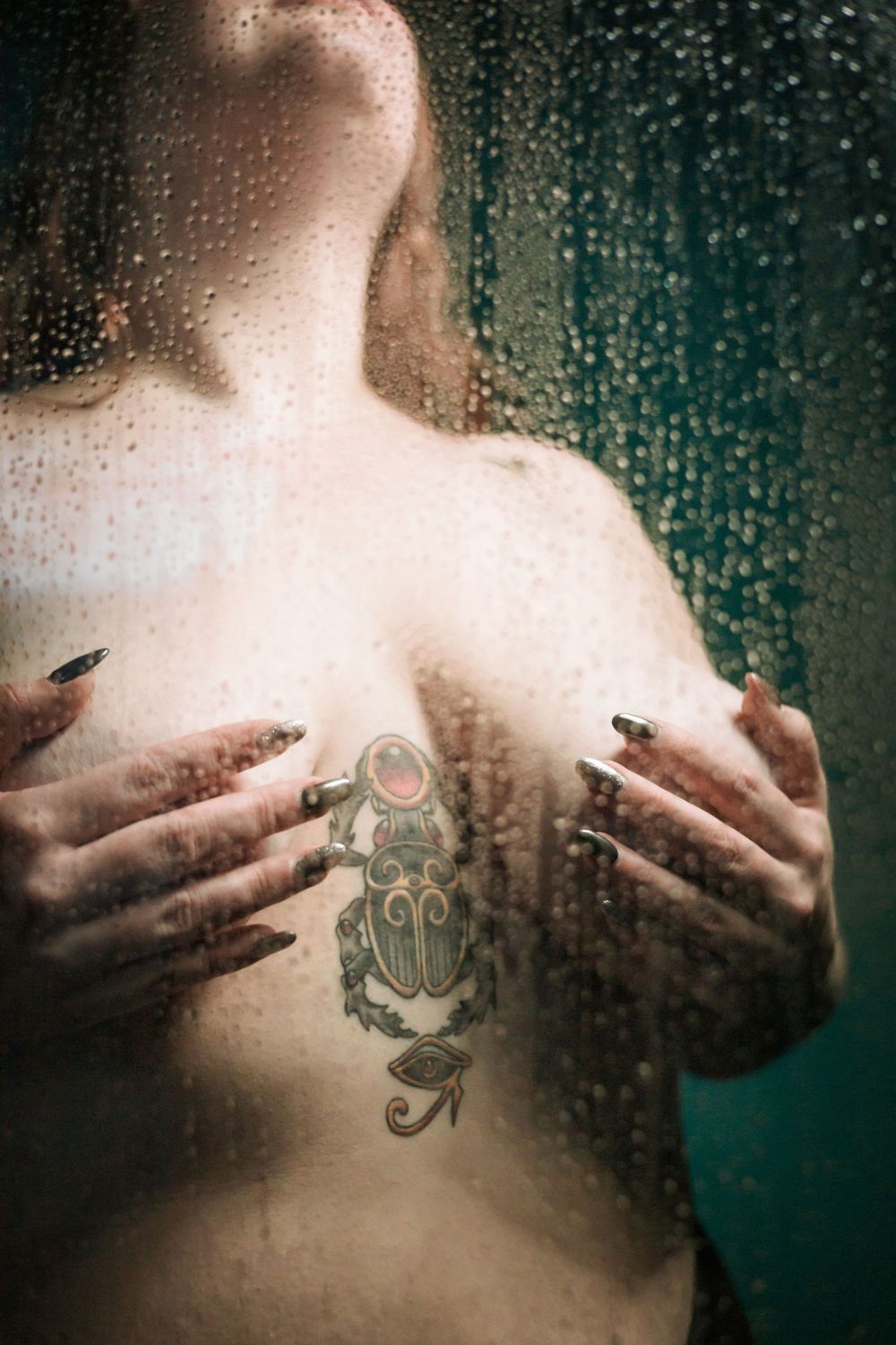 Artistic nude showing scarab tattoo behind steamy glass with water droplets.