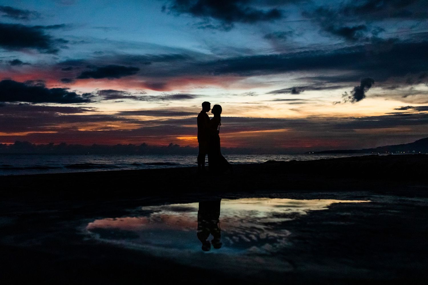A silhouetted couple stands reflected in water against a dramatic sunset sky.