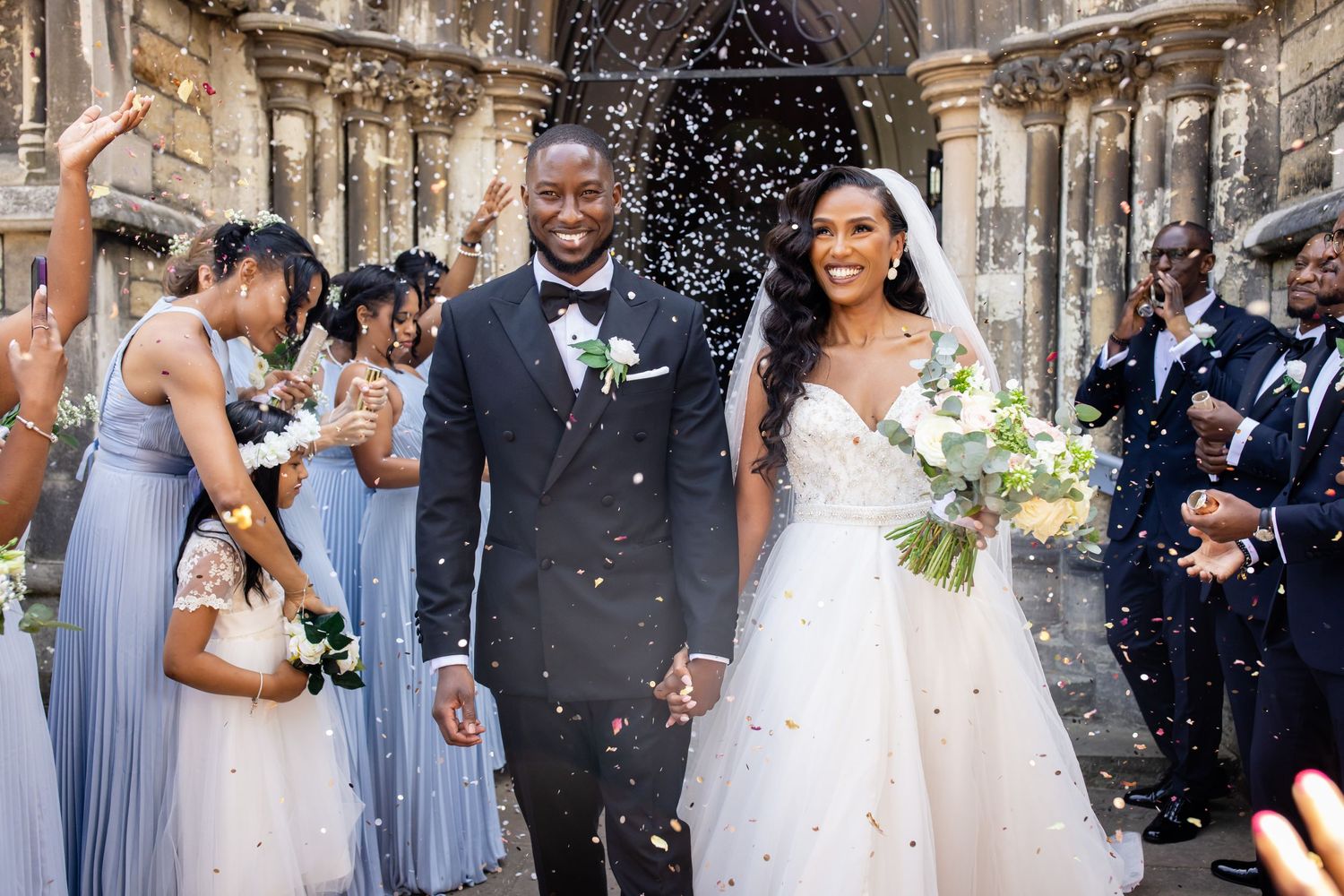 Wedding party celebrates with confetti as couple exits church in formal attire.