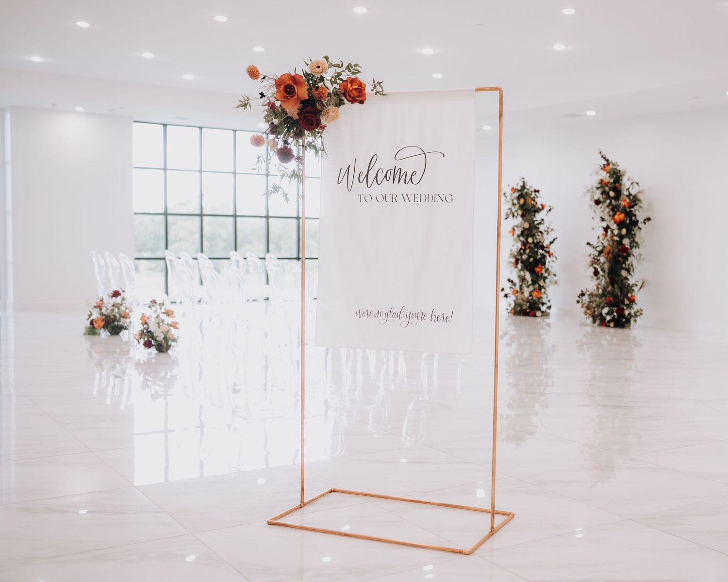 Modern wedding welcome sign with copper frame and floral arrangements in white minimalist space.