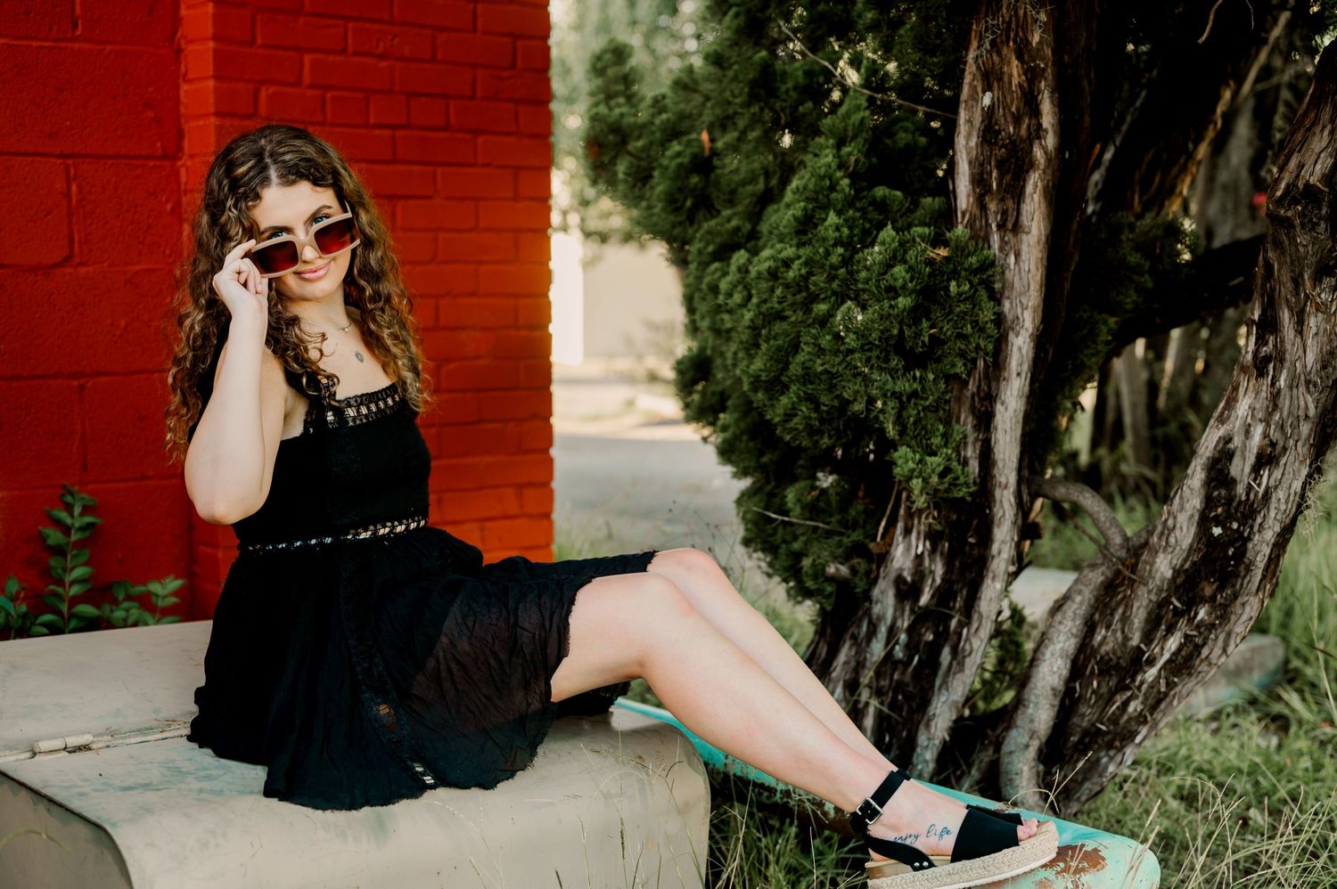 A fashion portrait against a red brick wall featuring a black dress and sunglasses in a casual outdoor setting.