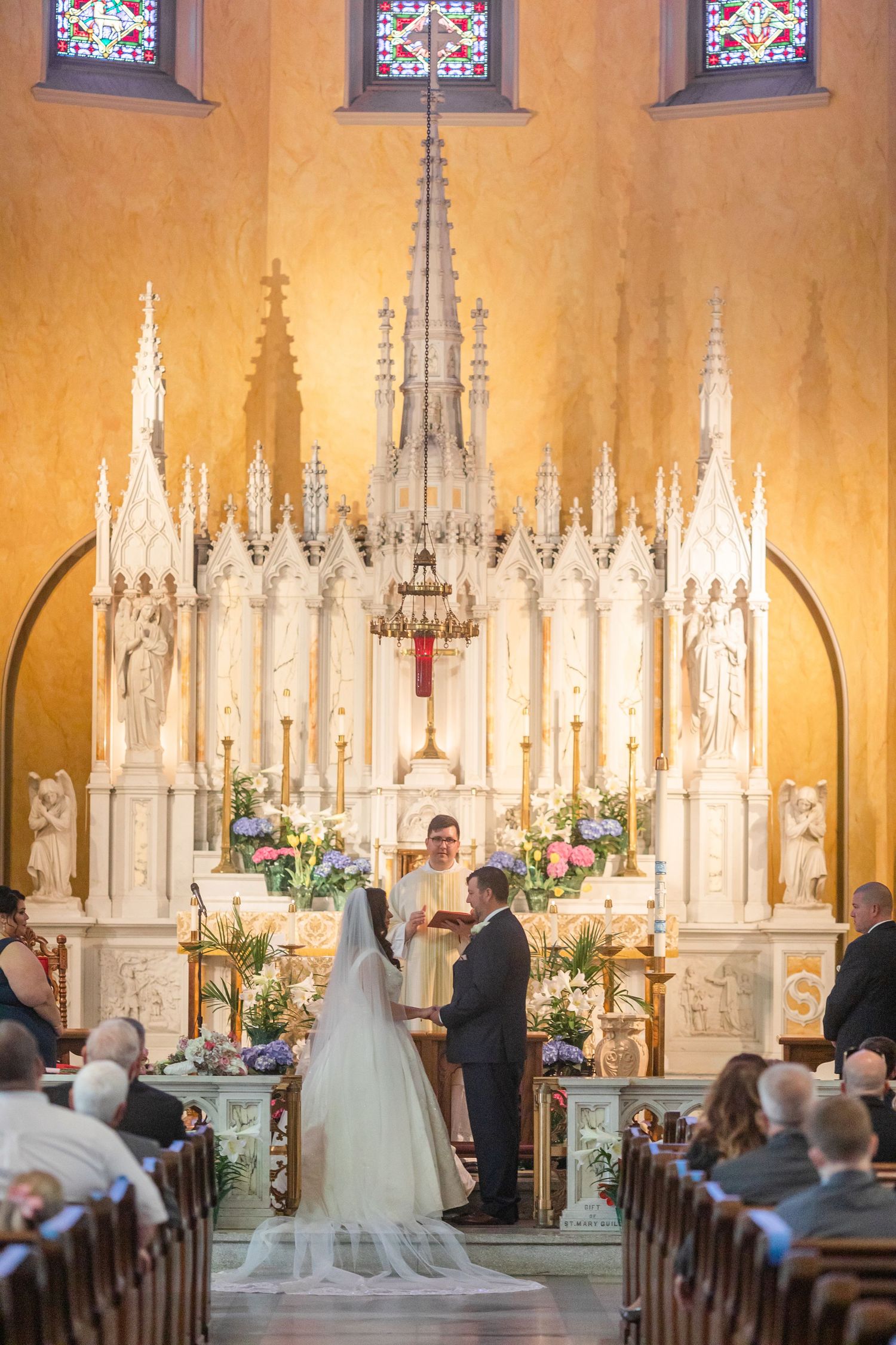 Cathedral wedding ceremony with intricate white altar and stained glass windows in warm lighting.