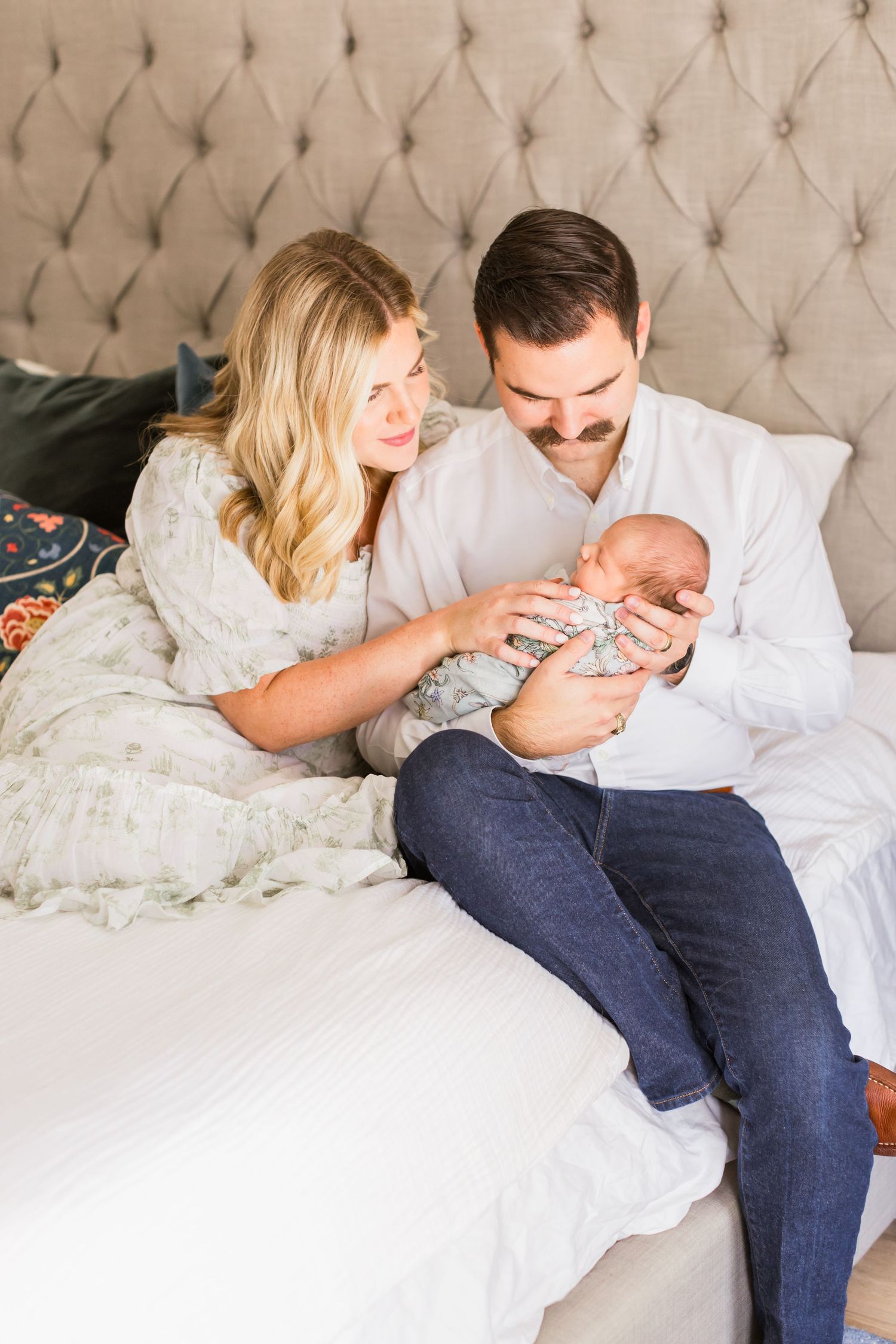 Parents sitting on bed together cradling their sleeping newborn.