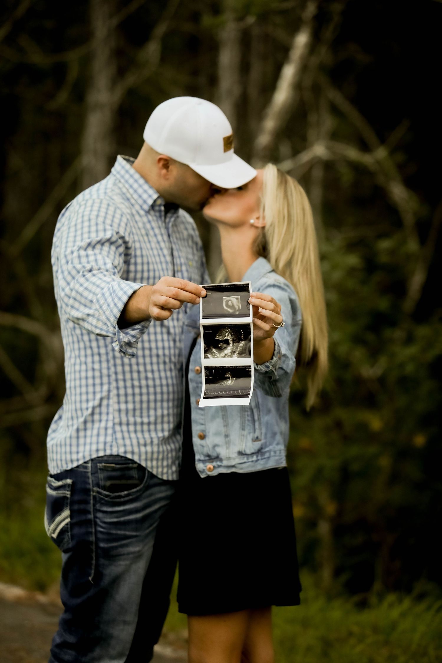 A couple shares an intimate moment while holding ultrasound photos in an outdoor setting.