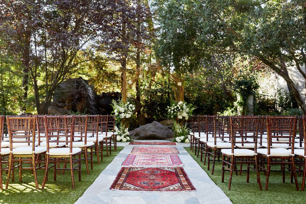 Outdoor wedding ceremony setup with wooden chairs and a Persian rug aisle runner under trees.
