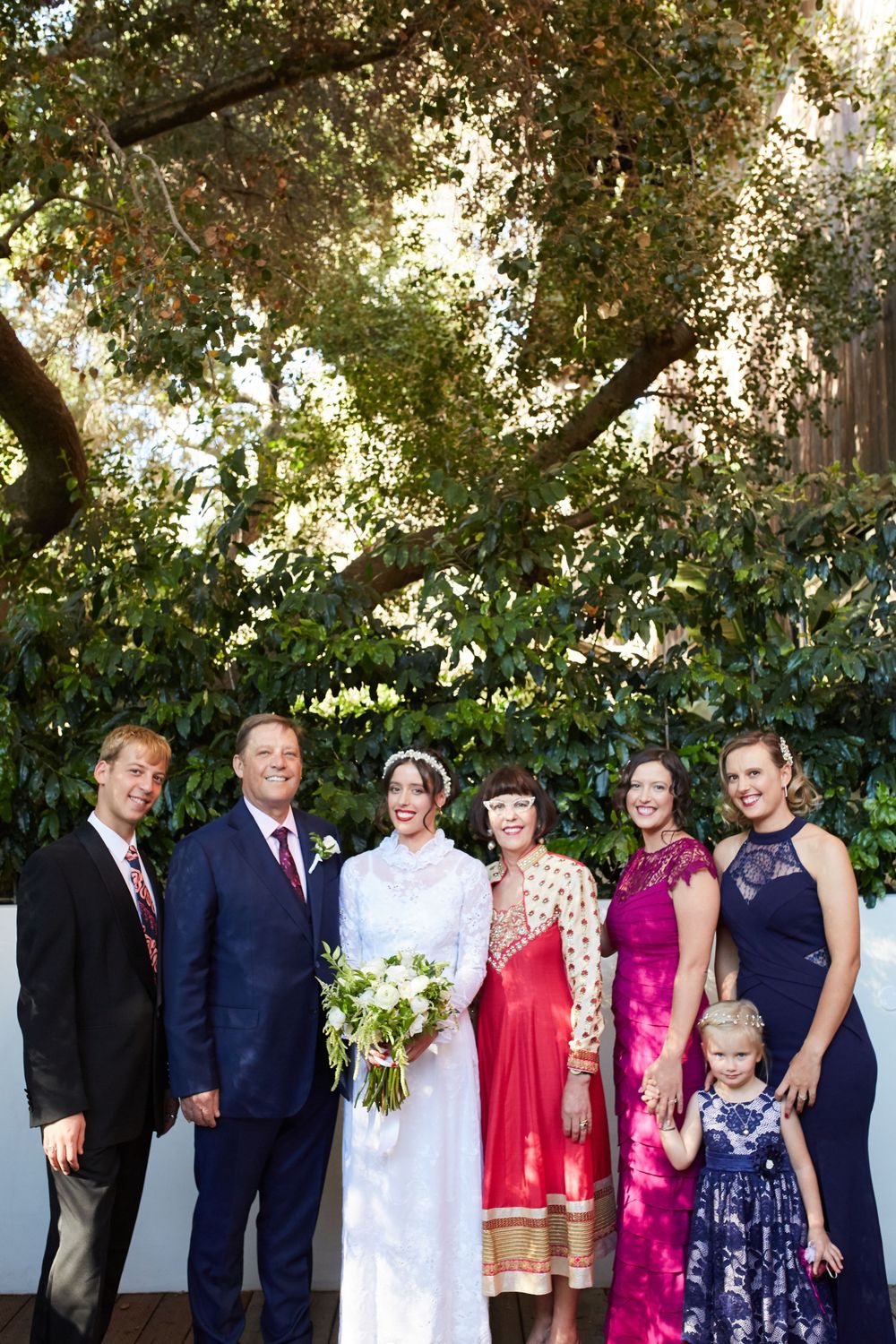 Wedding party standing together outdoors under lush trees with formal attire and floral bouquets.