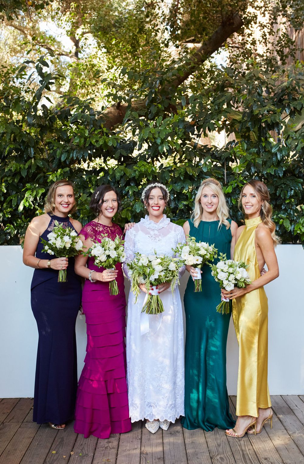 Bridal party in jewel-toned dresses posing with bouquets against a garden backdrop.