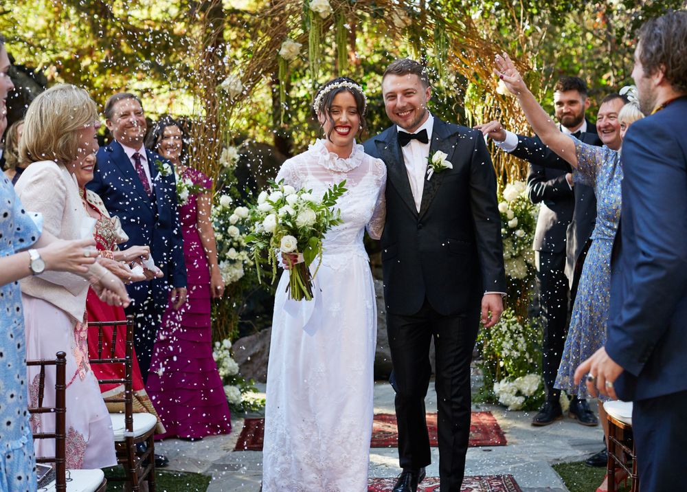 Wedding couple walking down the aisle while guests throw confetti in an outdoor garden setting.