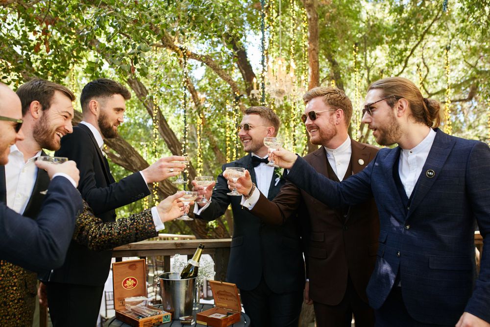 Groomsmen toasting with drinks at an outdoor wedding celebration under trees.