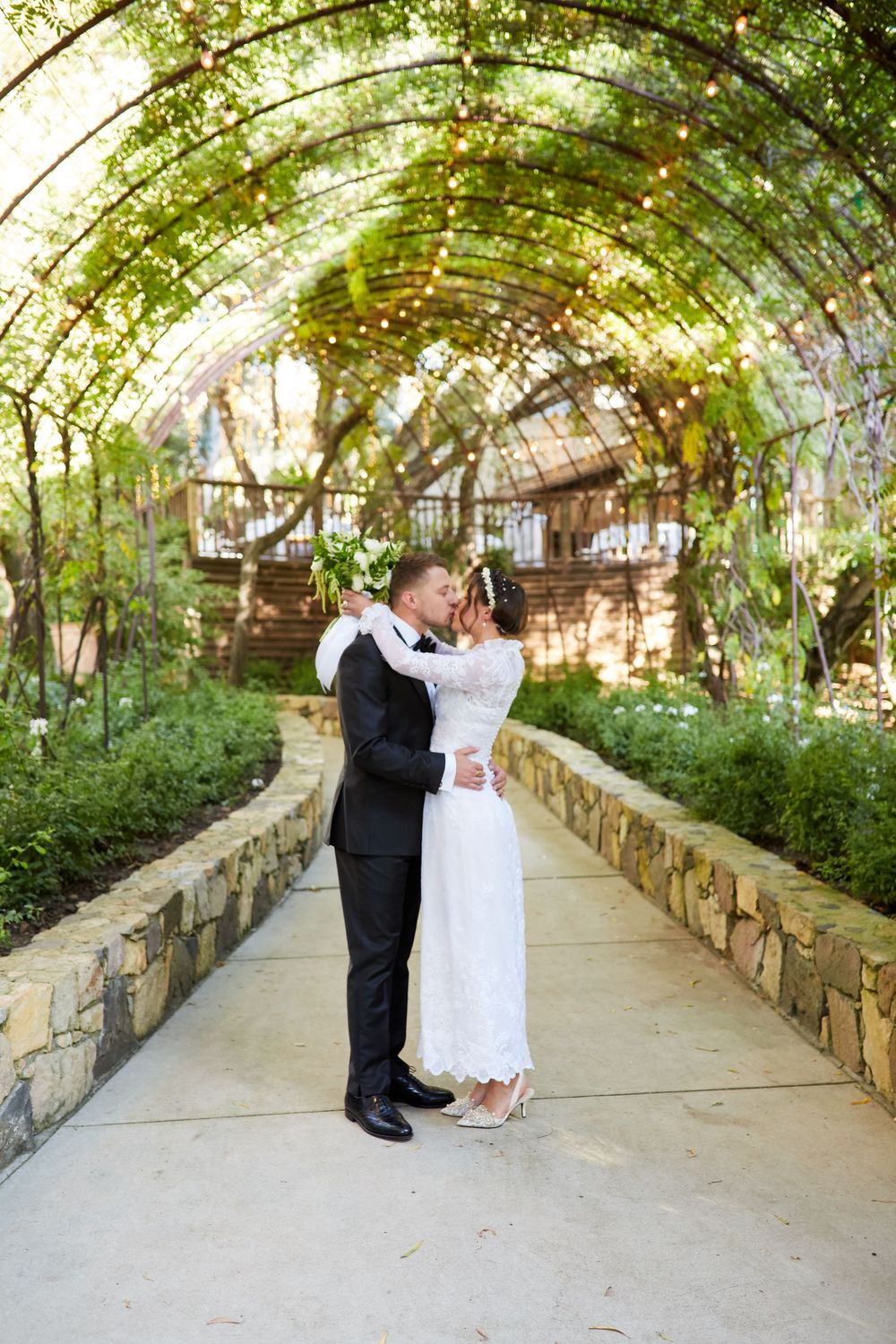 A romantic wedding scene under a vine-covered garden archway with stone walls.