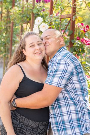 husband hugs and nuzzles his wife lovingly while standing under a rose archway.  Love.