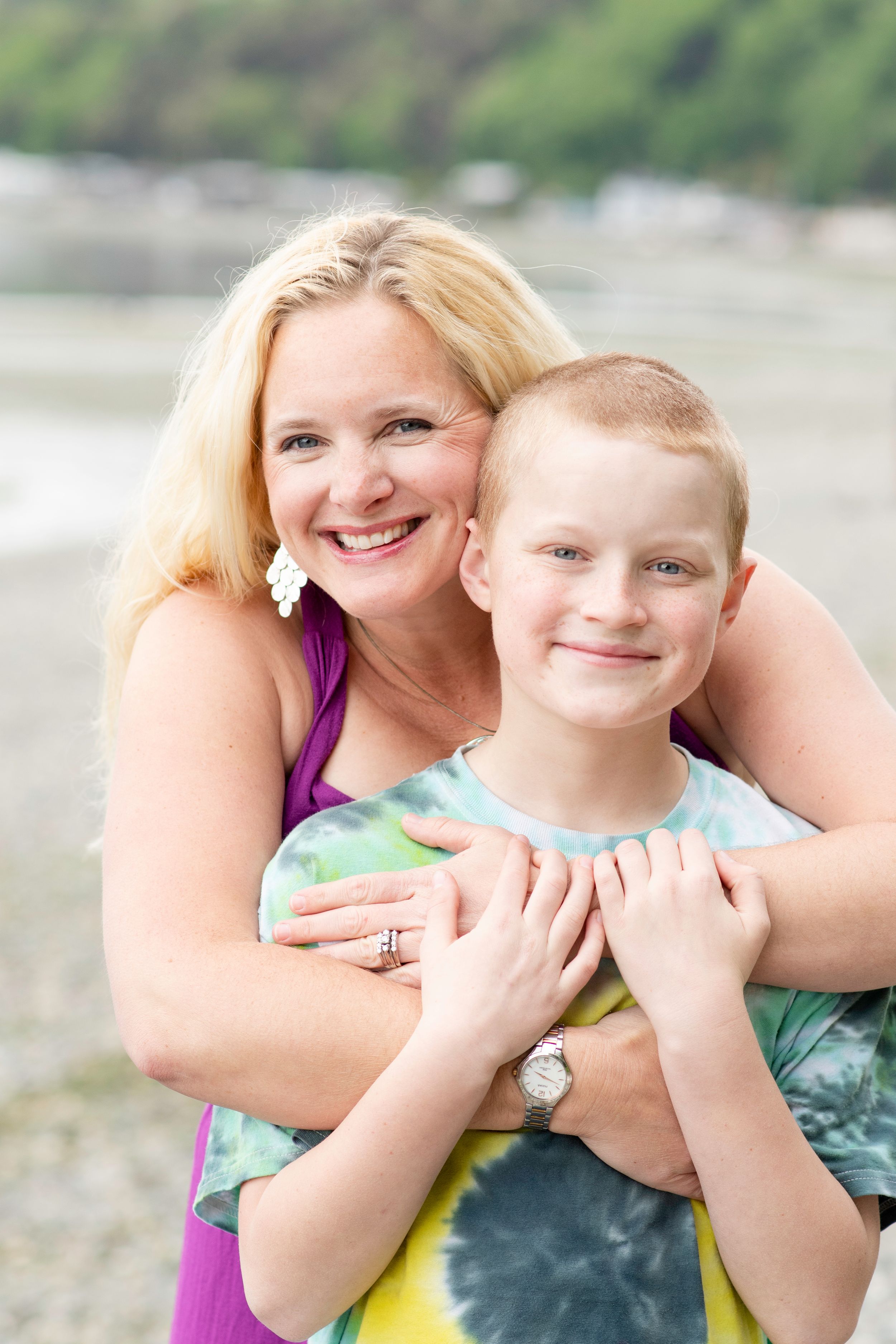 mother hugs her son from behind while standing on the beach shore framed by water and green trees.