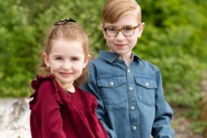 little sister looks in camera with smile and sits with big bother. They are framed with green trees.