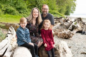 Family of four sits on driftwood logs at the beach. They are all smiling at the camera and framed by shore and trees.
