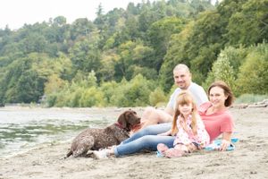 parents sit on the beach with their daughter and dog. They are relaxed in the sand and framed by green trees and water.