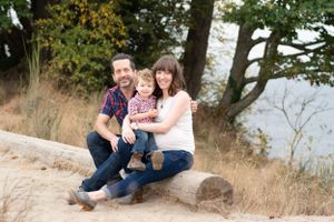 Pregnant mom holds toddler son and sits with husband on log with tall grass and trees framing them as well as the water.