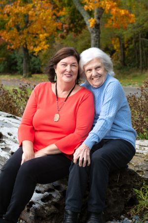 Silver haired mom snuggles her adult daughter as they laugh. Sitting framed by popping fall leaves.