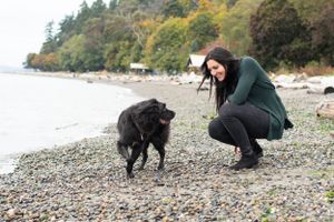 Woman squats down to lovingly talk to her dog who is prancing along the water shoreline.