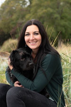 woman holds her black dog close while sitting in the green beach grass. Her eyes are green and she is framed by trees.