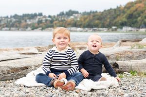 baby and toddler brother sit on a blanket on the rocky PNW beach and laugh. Framed by the tree lined shore.