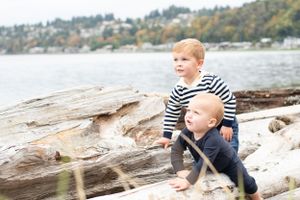 baby and his toddler brother look curiously into the distance while leaning on driftwood logs at the beach.