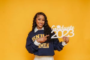 Smiling graduate in SNHImen sweatshirt pointing at Graduate 2023 sign on yellow background.