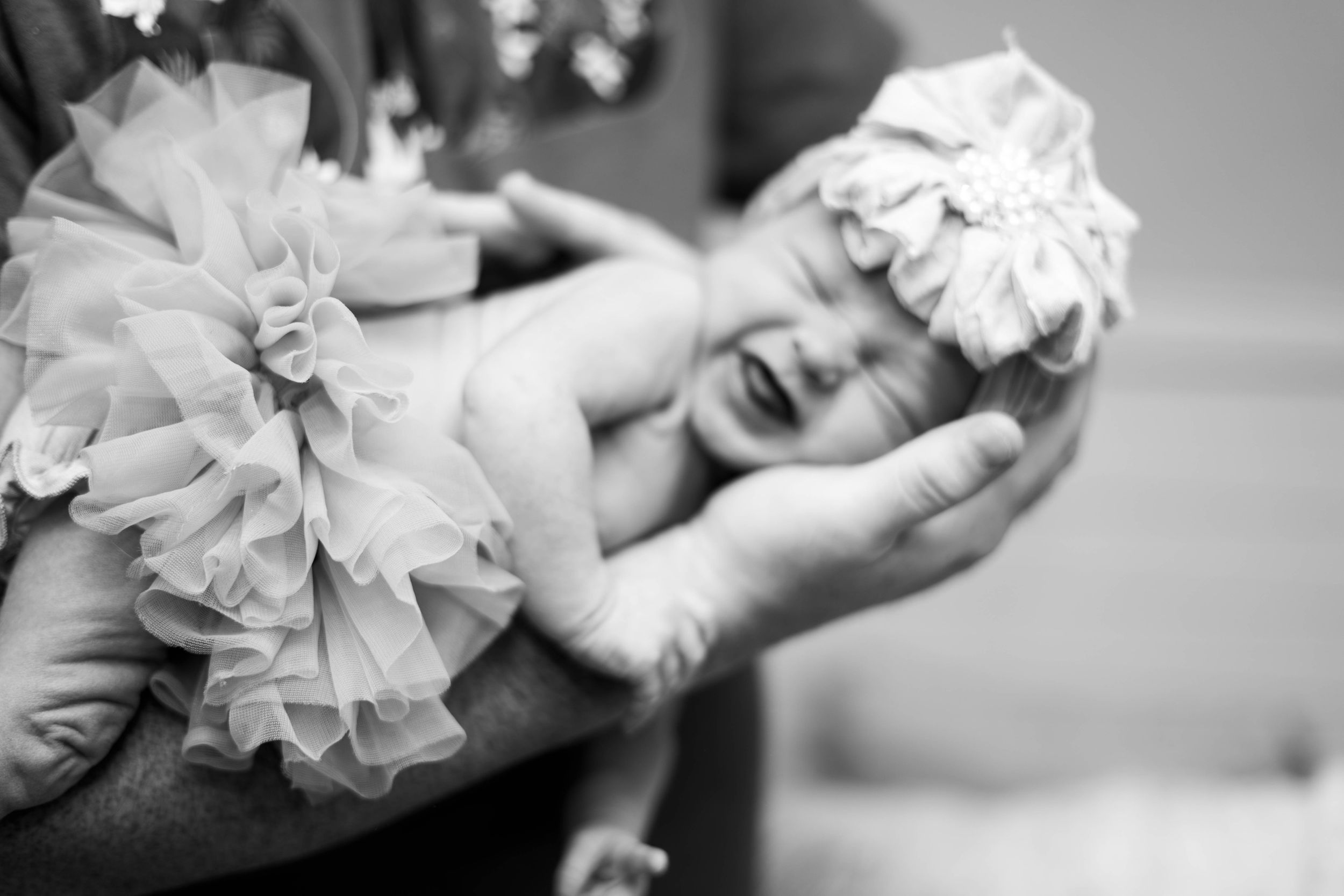 Black and white photo of a crying newborn baby in a tutu dress being cradled in loving hands.