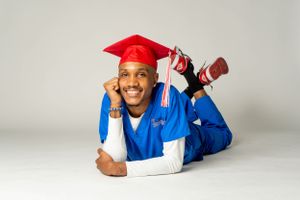 Proud graduate in red cap and blue gown lying down smiling against a clean white studio background.