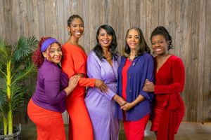 Five stylish women in vibrant red and purple outfits smiling together in front of a wooden backdrop.
