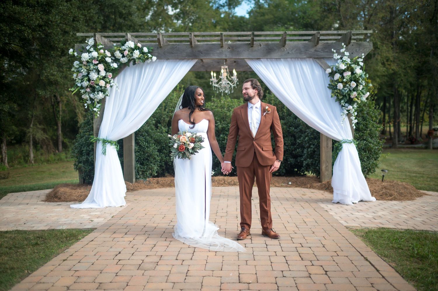 Wedding ceremony setup with white draped arch and floral arrangements on brick paved outdoor area.