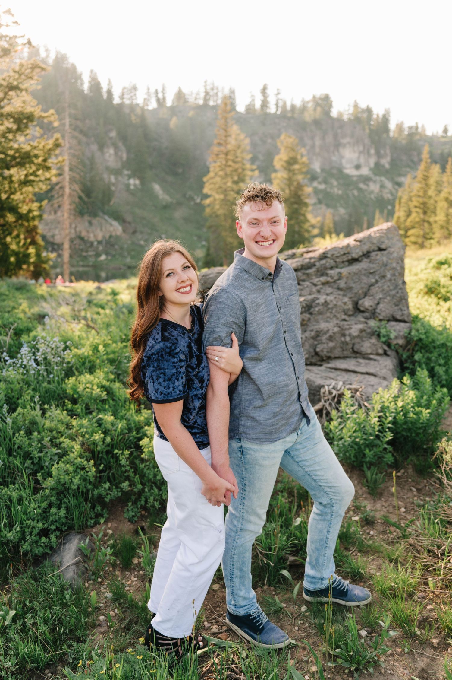 A couple poses together on a mountain trail surrounded by evergreen trees and rocky cliffs.