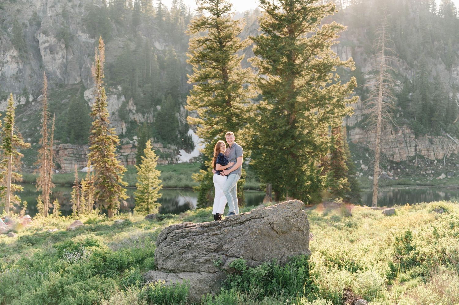 A couple stands on a large boulder with mountains and pine trees in the background.