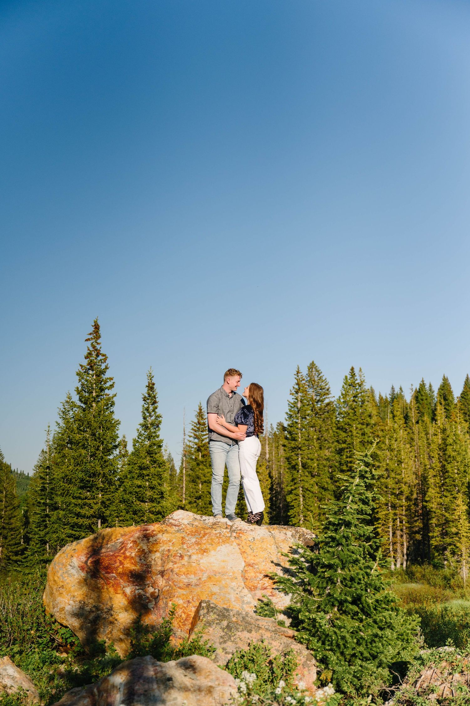 A couple stands atop a large boulder with evergreen trees and blue sky in the background.