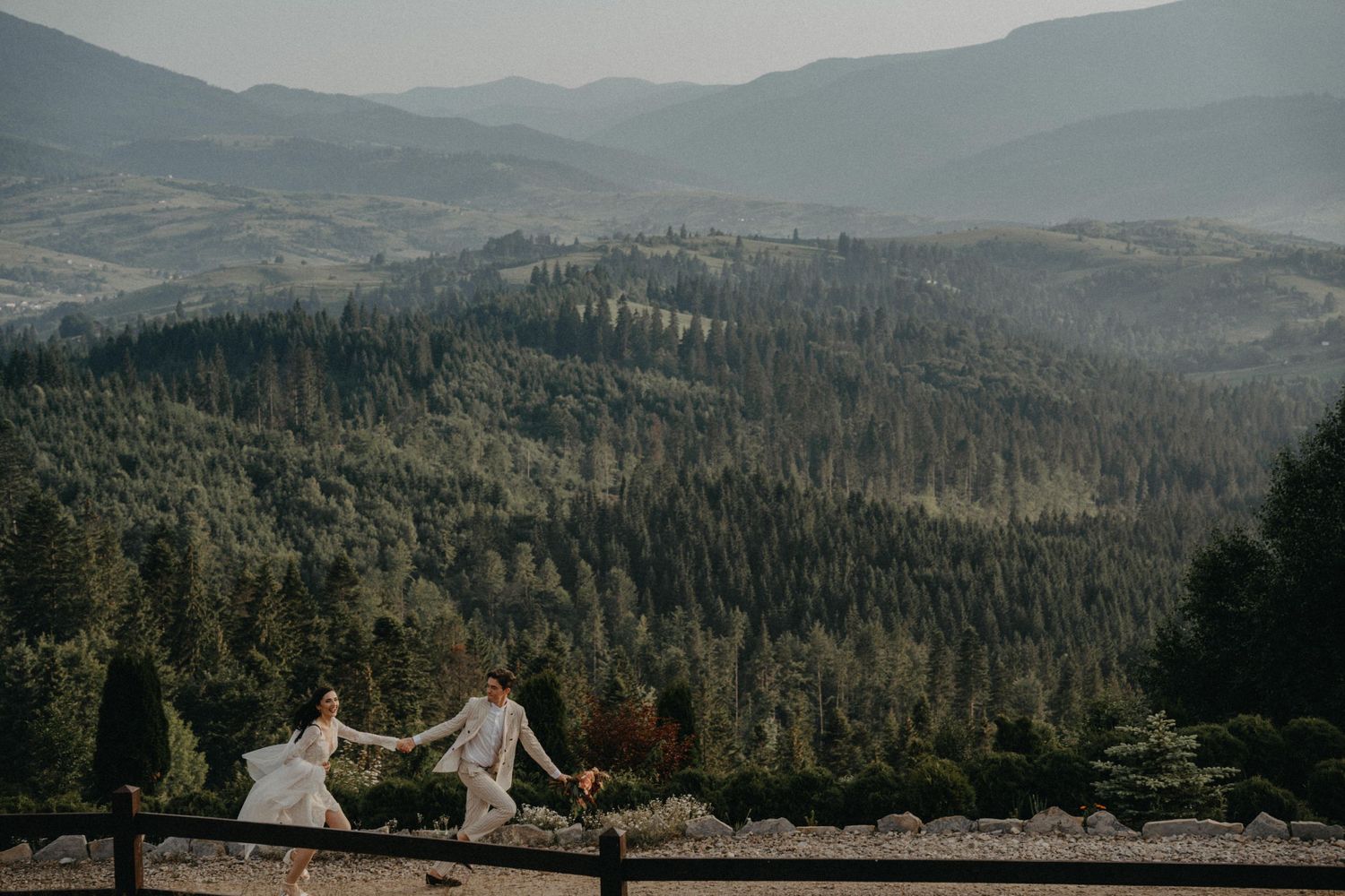 Couple holding hands on a mountain overlook surrounded by lush pine forest and misty valley views.