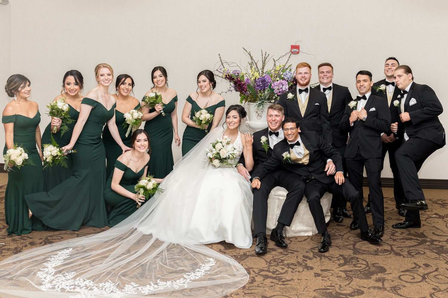 Wedding party poses together with bridesmaids in emerald green dresses and groomsmen in black tuxedos.