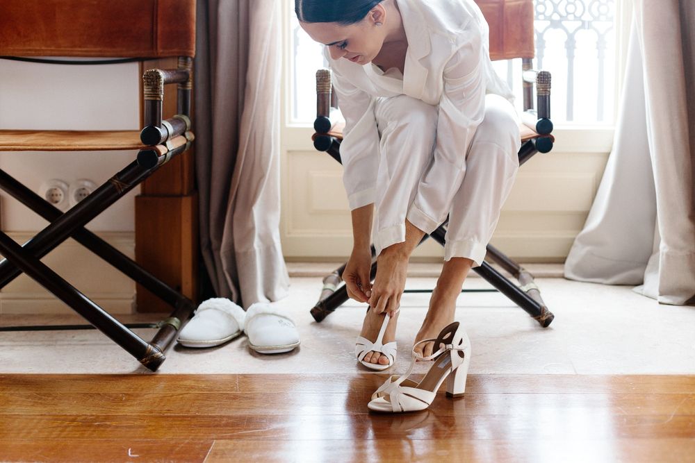 Close-up of someone in white putting on elegant bridal shoes while seated.