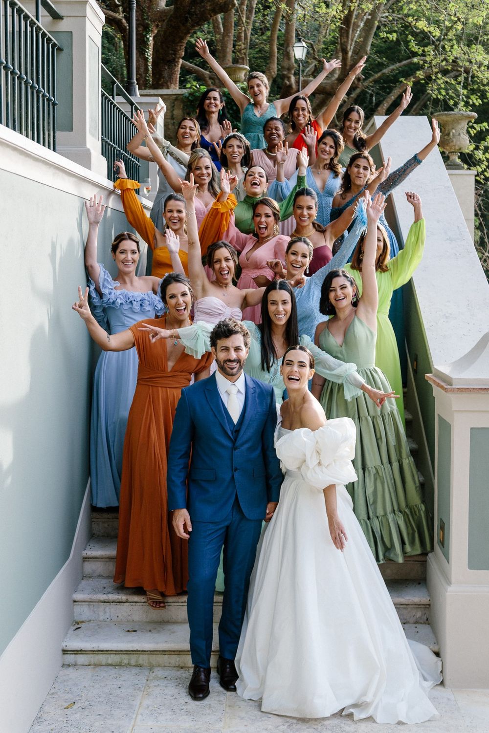Wedding party in colorful dresses poses joyfully on white steps with newlyweds in blue suit and white gown.