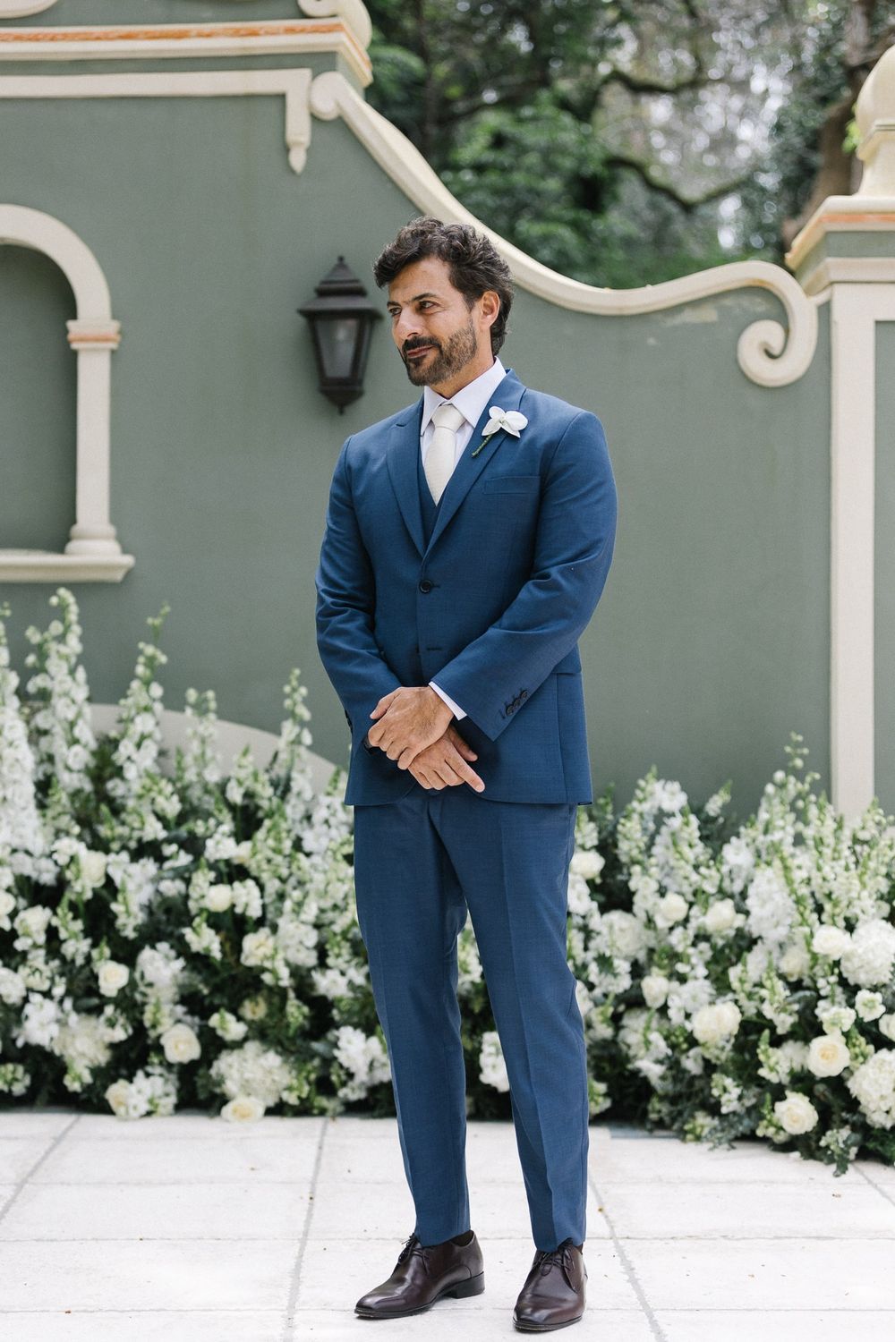 Groom in tailored navy blue suit standing against floral garden wall.