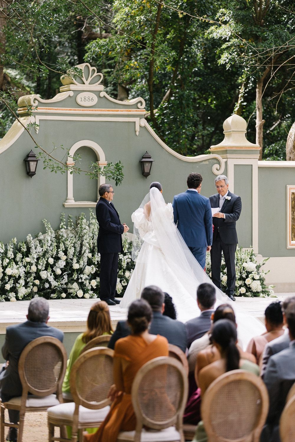 An outdoor wedding ceremony with white floral decorations against a gray Spanish-style wall.