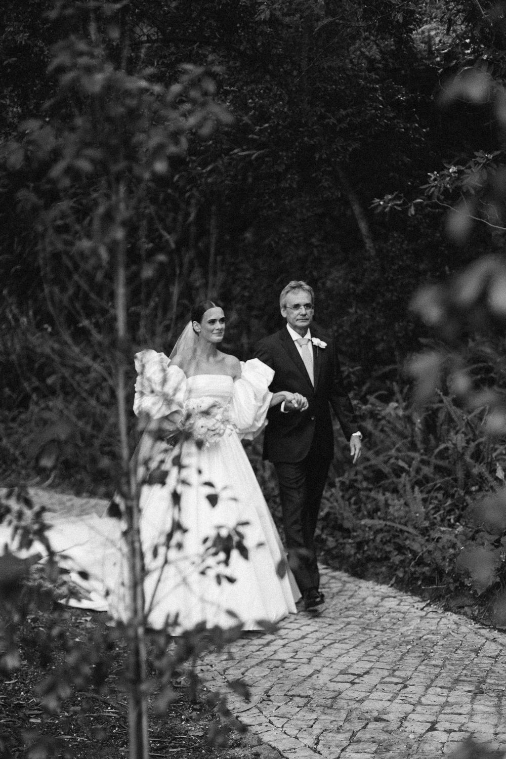 A black and white photo captures a wedding day entrance along a stone path surrounded by greenery.