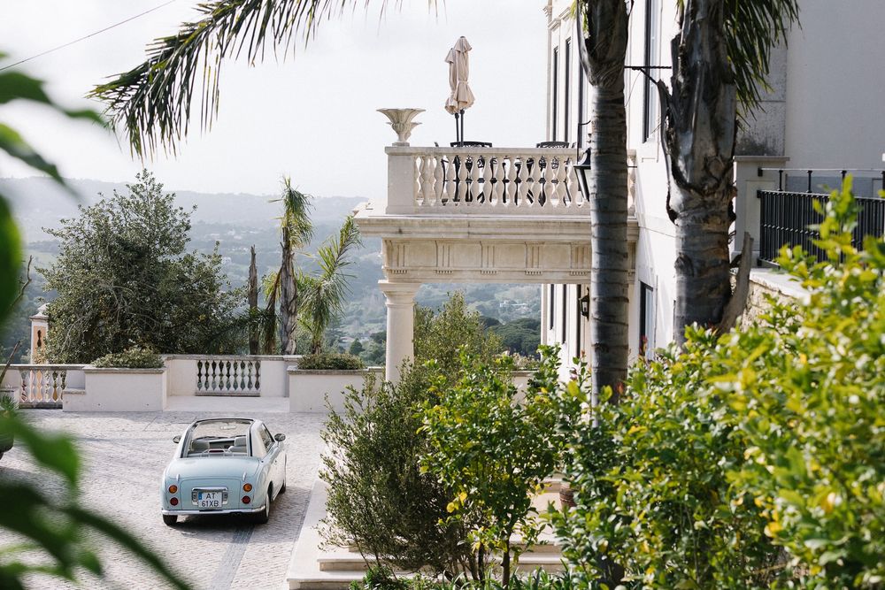 Classic car parked outside Mediterranean villa with palm trees and ornate balcony.