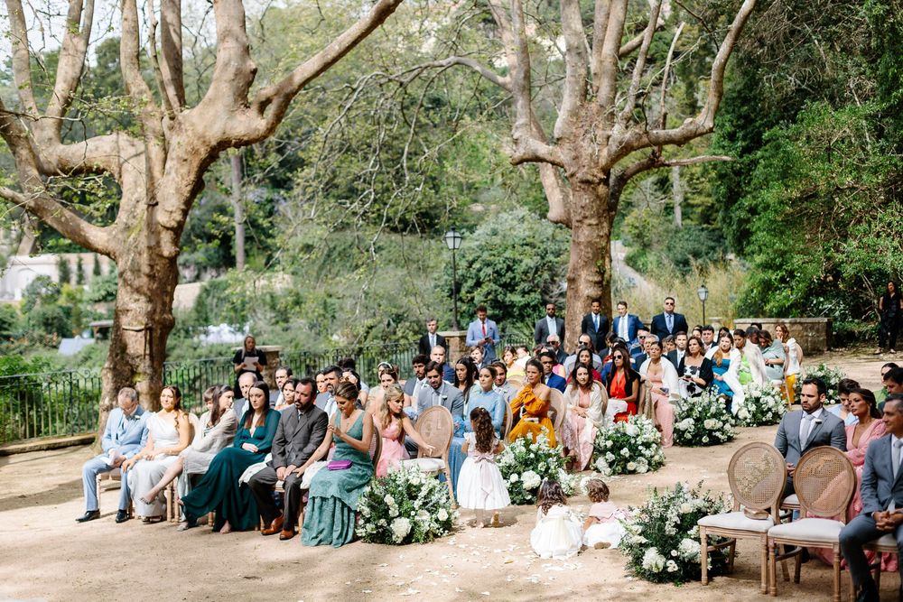 Garden wedding ceremony with guests seated among trees and floral arrangements.
