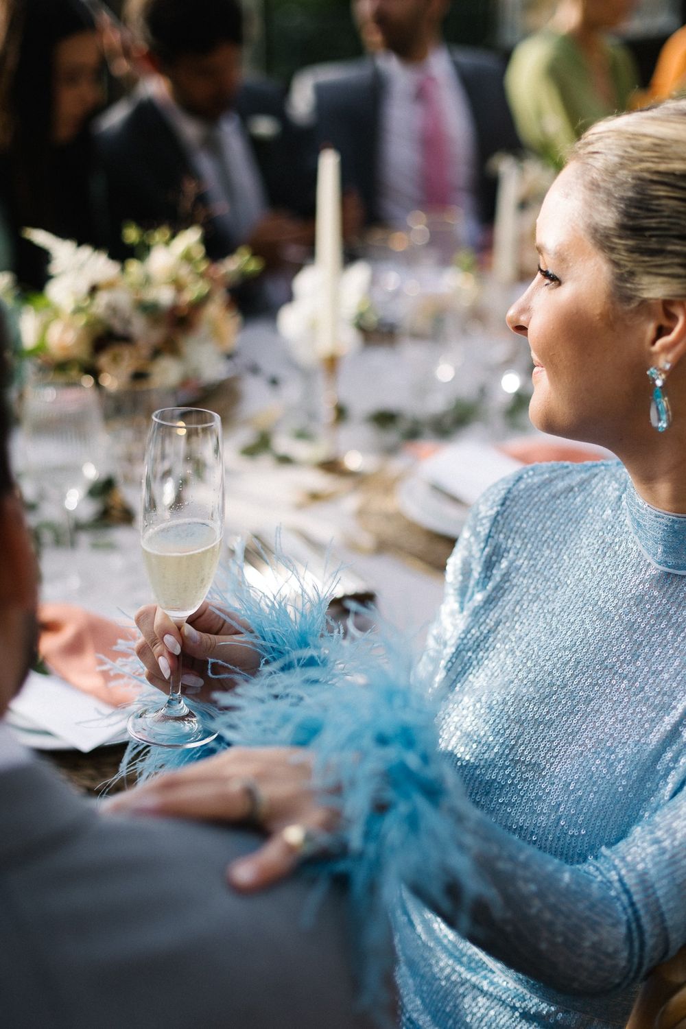 Guest in sparkling blue dress with feather detail enjoys champagne at wedding reception.