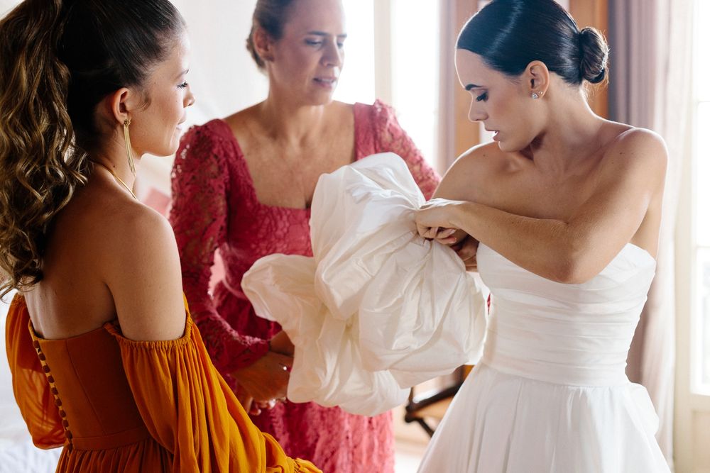 Women in colorful dresses helping adjust a white wedding gown during preparation.