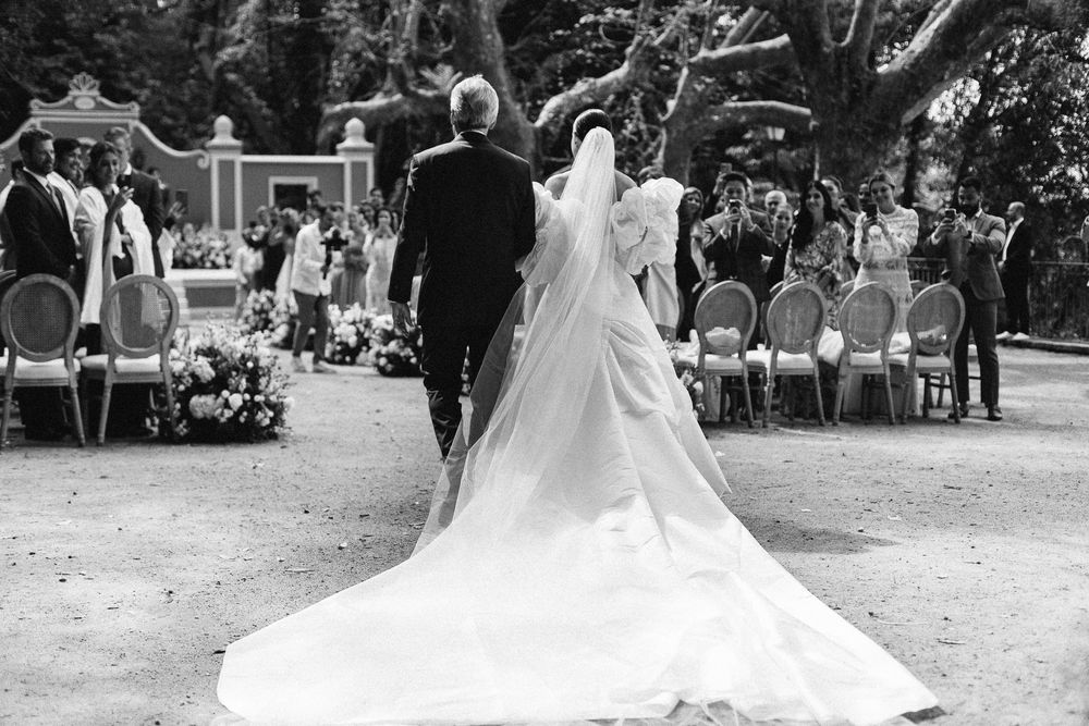 A stunning black and white wedding ceremony photo shows a trailing white dress on a garden path with guests seated.