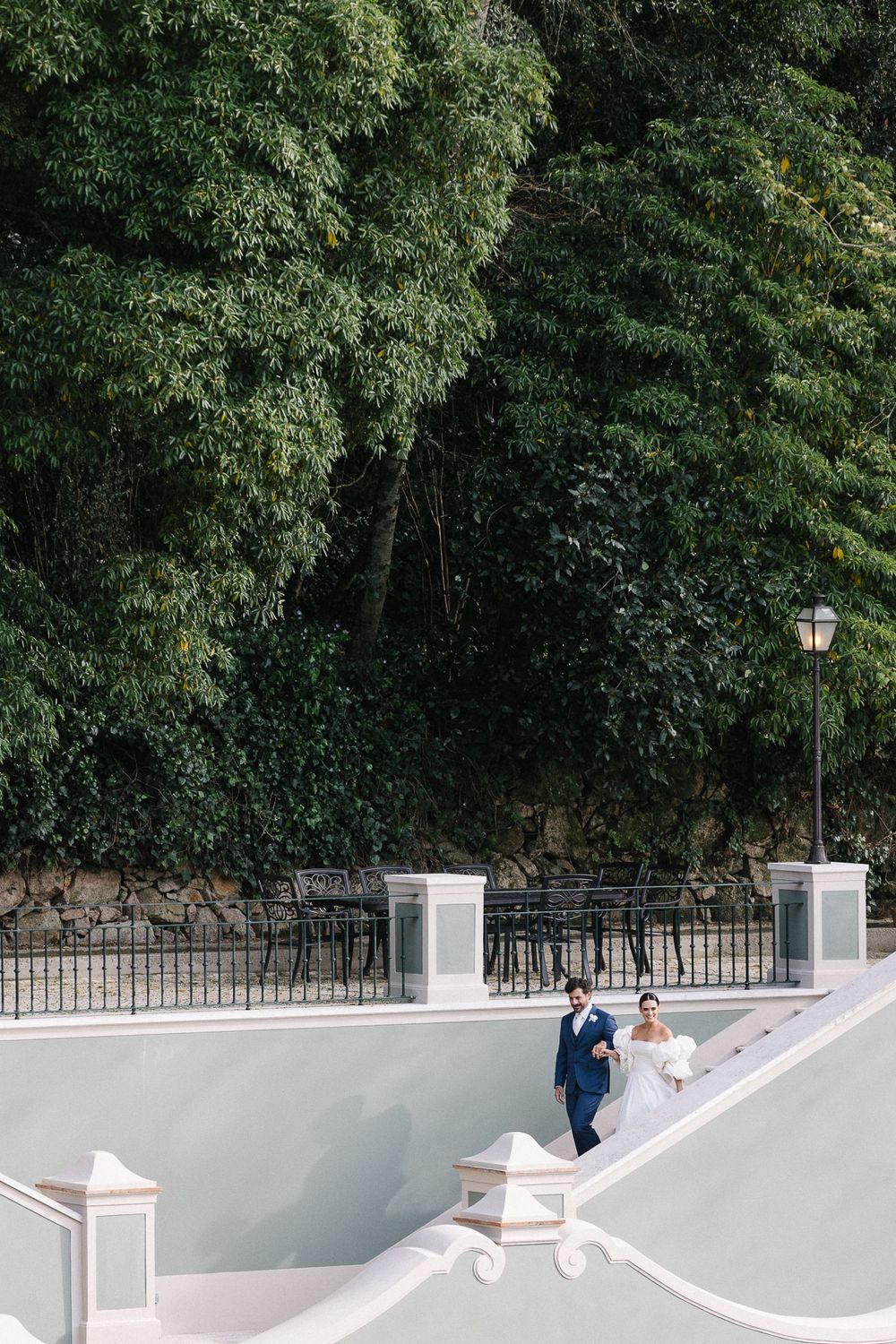 Wedding couple walking down white staircase surrounded by lush greenery and iron railings.