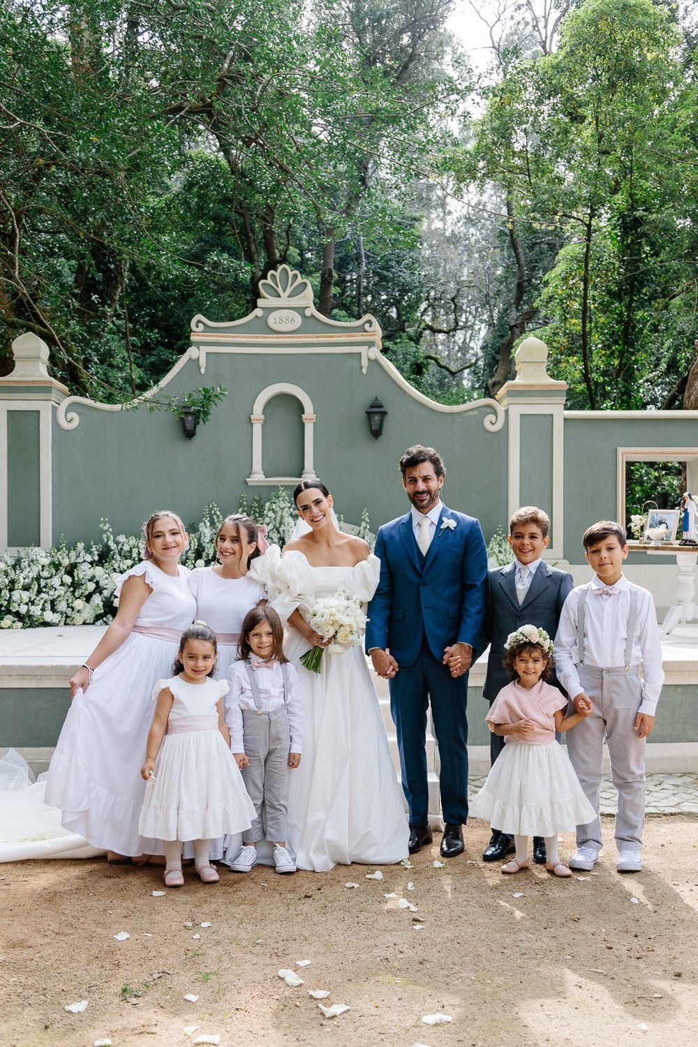 Wedding party poses in front of a decorative garden wall with children in white formal attire.