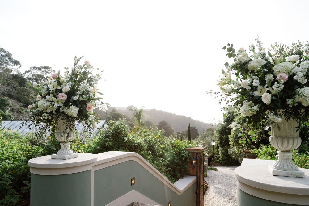 White floral arrangements in stone urns frame a scenic outdoor wedding venue view.
