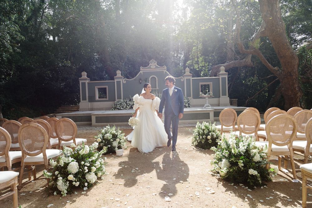 Outdoor wedding ceremony setup with wooden chairs and floral arrangements framing a decorative wall.